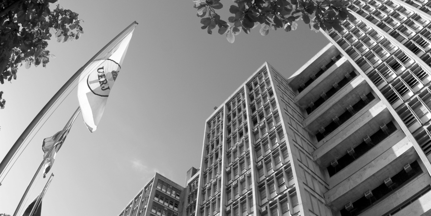 Fotografia externa, em preto e branco, do Campus Maracanã. À esquerda, a bandeira hasteada da Uerj e, à direita, o prédio da universidade.
